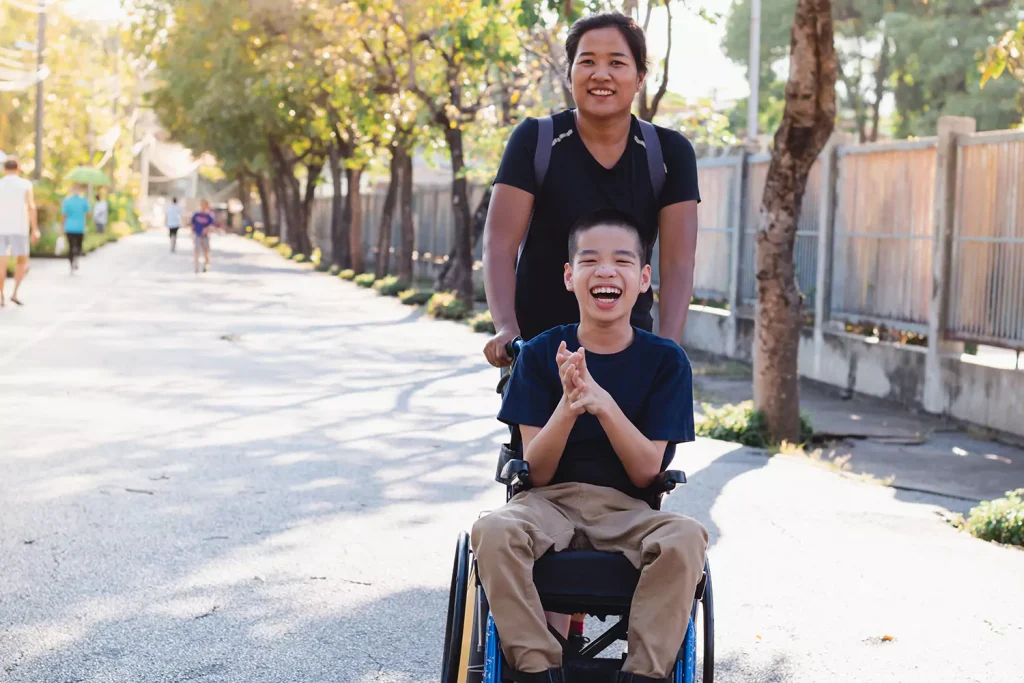 A disabled boy in his wheelchair while his support worker walks him in the park, happily enjoying disability support